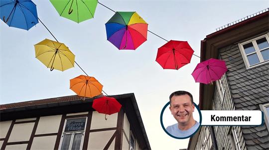 Bunte Regenschirme hängen über einem Fachwerkhaus, rechts ein Mann mit kurzem Haar und blauem Hemd lächelt, daneben das Wort 'Kommentar'