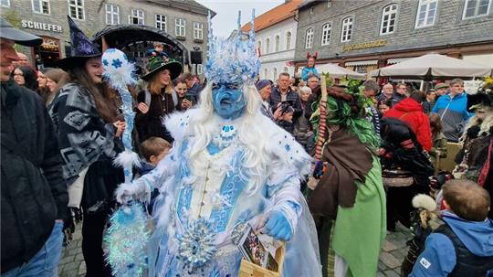 Menschenmenge auf einem Marktplatz mit zwei Personen in auffälligen Kostümen, eine in blau-weißer Verkleidung mit langem weißen Haar und eine in grüner Verkleidung mit Kopfschmuck aus Blättern
