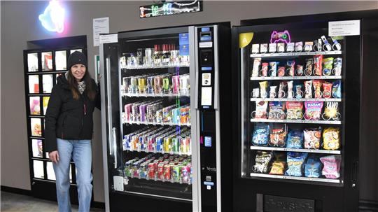 Person steht neben drei Verkaufsautomaten mit Getränken und Snacks in einem Innenraum.