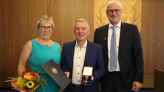 Drei Personen in formeller Kleidung stehen nebeneinander, die Frau links hält einen Blumenstrauß, der Mann in der Mitte zeigt eine Medaille in einer offenen Schachtel