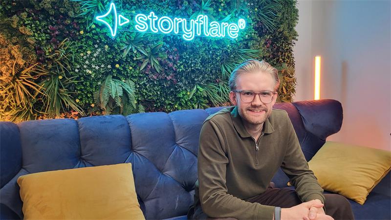 Mann mit Brille und Pullover sitzt auf blauem Sofa vor Wand mit Pflanzen und leuchtendem Schriftzug 'storyflare'