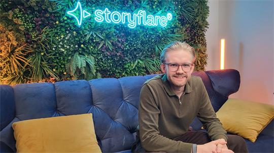 Mann mit Brille und Pullover sitzt auf blauem Sofa vor Wand mit Pflanzen und leuchtendem Schriftzug 'storyflare'