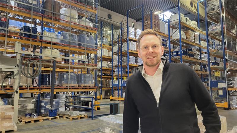 Mann in schwarzer Jacke steht in einem Lager mit hohen Regalen voller Kartons und Behälter