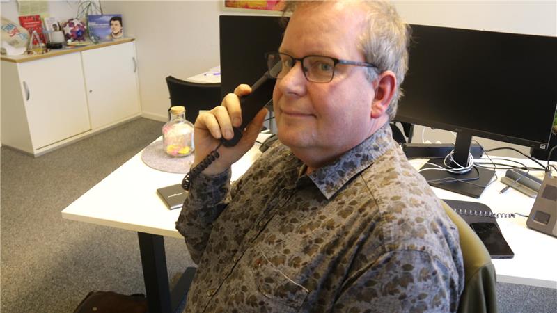 Person mit gemustertem Hemd sitzt an einem Schreibtisch und hält einen Telefonhörer ans Ohr.