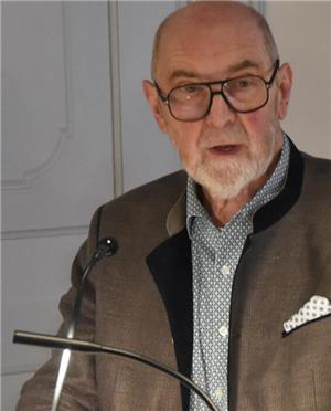 Älterer Mann mit grauem Sakko, gemustertem Hemd und Einstecktuch steht vor einem Mikrofon in einem Raum mit weißer Tür im Hintergrund