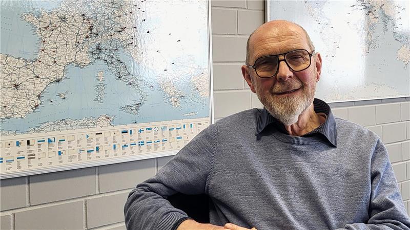 Mann mit grauem Pullover sitzt vor zwei Landkarten an einer weißen Kachelwand.