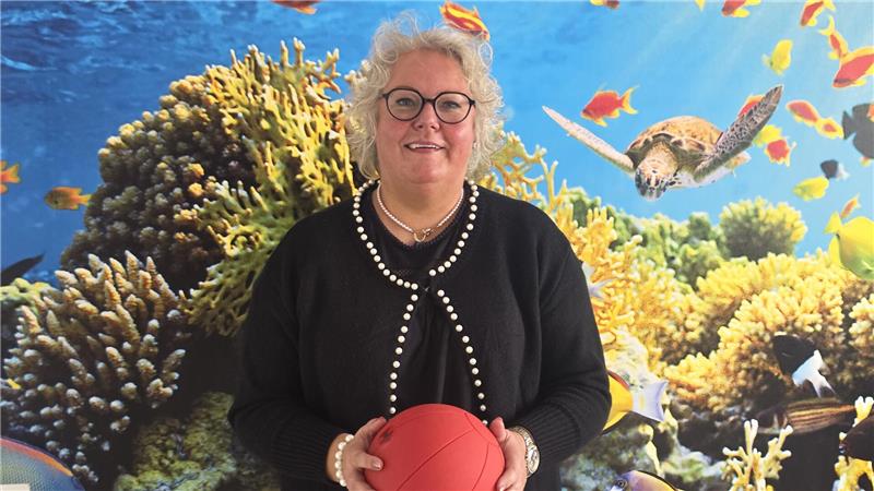 Person mit schwarzem Pullover und Perlenkette hält roten Ball vor einer Wand mit Unterwasser-Szene mit Korallen, Fischen und einer Schildkröte.