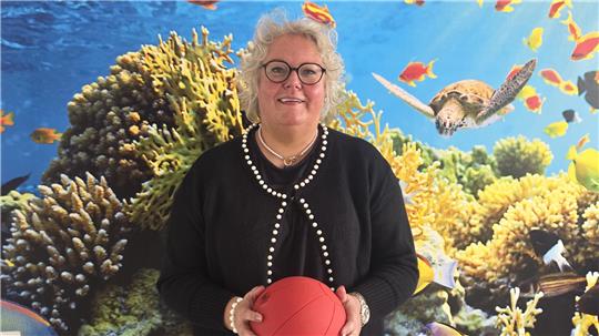 Person mit schwarzem Pullover und Perlenkette hält roten Ball vor einer Wand mit Unterwasser-Szene mit Korallen, Fischen und einer Schildkröte.