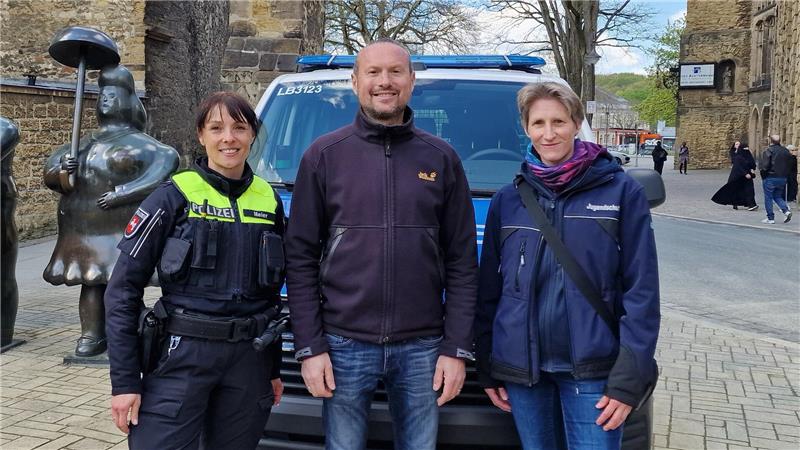 Drei Personen stehen vor einem Polizeifahrzeug auf einem gepflasterten Platz mit historischen Gebäuden im Hintergrund