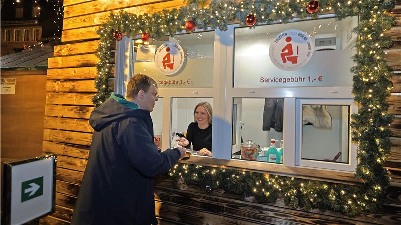 GZ-Redakteur Sowa im WC-Einsatz: Zwischen Klobürste und Flatrate Mann in dunklem Mantel steht an einem weihnachtlich geschmückten WC-Kassen-Holzstand mit zwei Fenstern, an einem Fenster bedient eine Frau.