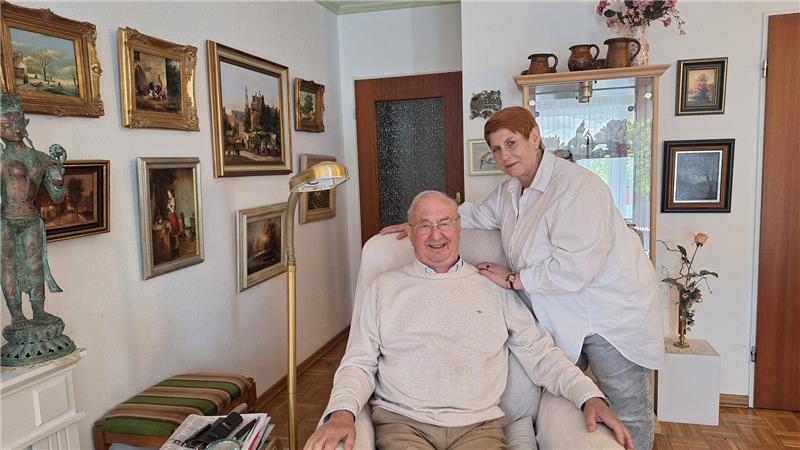 Ein älterer Mann sitzt in einem Sessel, während eine Frau daneben steht und ihn an den Schultern festhält. 