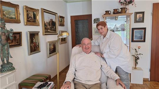 Ein älterer Mann sitzt in einem Sessel, während eine Frau daneben steht und ihn an den Schultern festhält. 