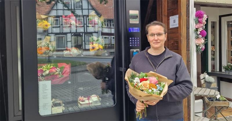 Person mit Brille und dunkler Jacke hält einen Blumenstrauß vor einem Geschäft mit Blumen und einem Schild 'Blütenzauber'