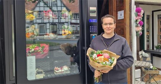 Person mit Brille und dunkler Jacke hält einen Blumenstrauß vor einem Geschäft mit Blumen und einem Schild 'Blütenzauber'