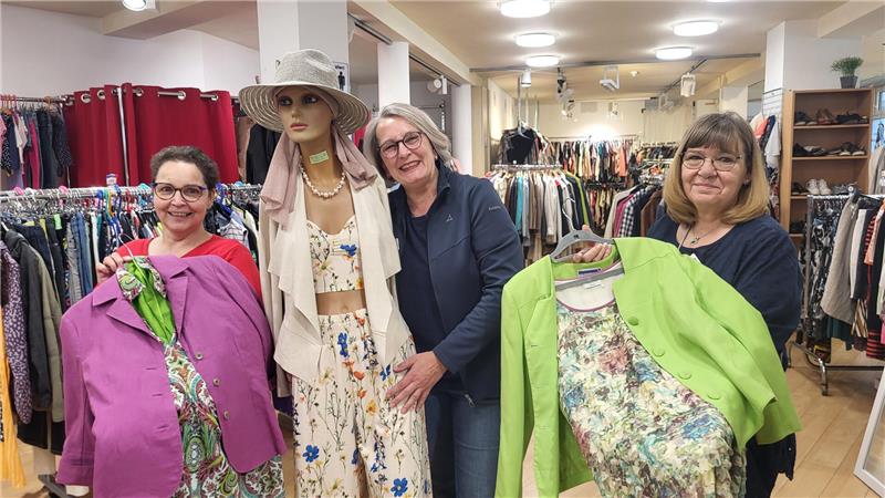 Drei Frauen in einem Bekleidungsgeschäft halten bunte Kleidungsstücke vor sich, im Hintergrund sind Kleiderständer mit weiteren Kleidern zu sehen.