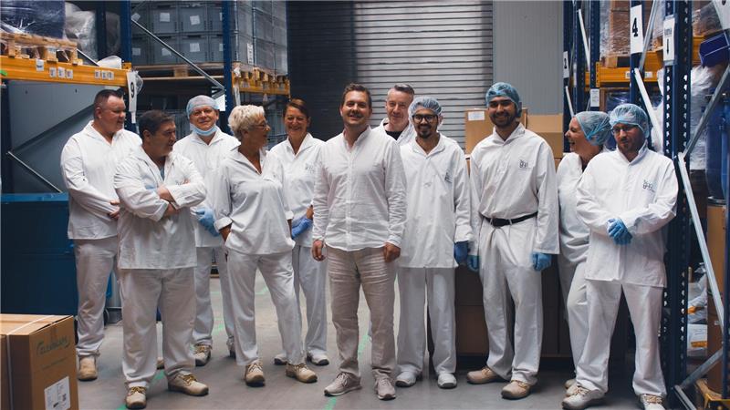 Menschen mit weißen Schutzanzügen, blauen Handschuhen und Haarnetzen stehen in einer Lagerhalle.