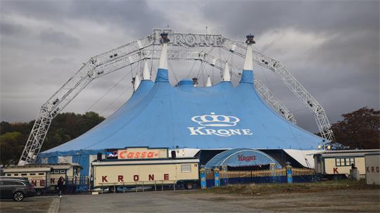 Das Bild zeigt den Circus Krone mit dem großen blauen Zirkuszelt, dem Eingangsbereich und mehreren Wohn- bzw. Betriebswagen. Im Hintergrund ist ein bewölkter Himmel zu sehen.