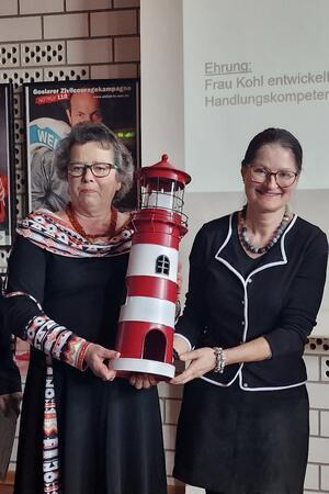 Zwei Frauen in schwarzer Kleidung halten gemeinsam ein rot-weiß gestreiftes Modell eines Leuchtturms