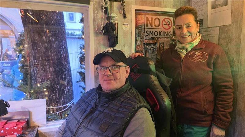 Zwei Personen in einem kleinen Raum mit Holzvertäfelung, eine sitzt in einem Gaming-Stuhl, die andere steht daneben. An der Wand hängt ein "No Smoking"-Schild.