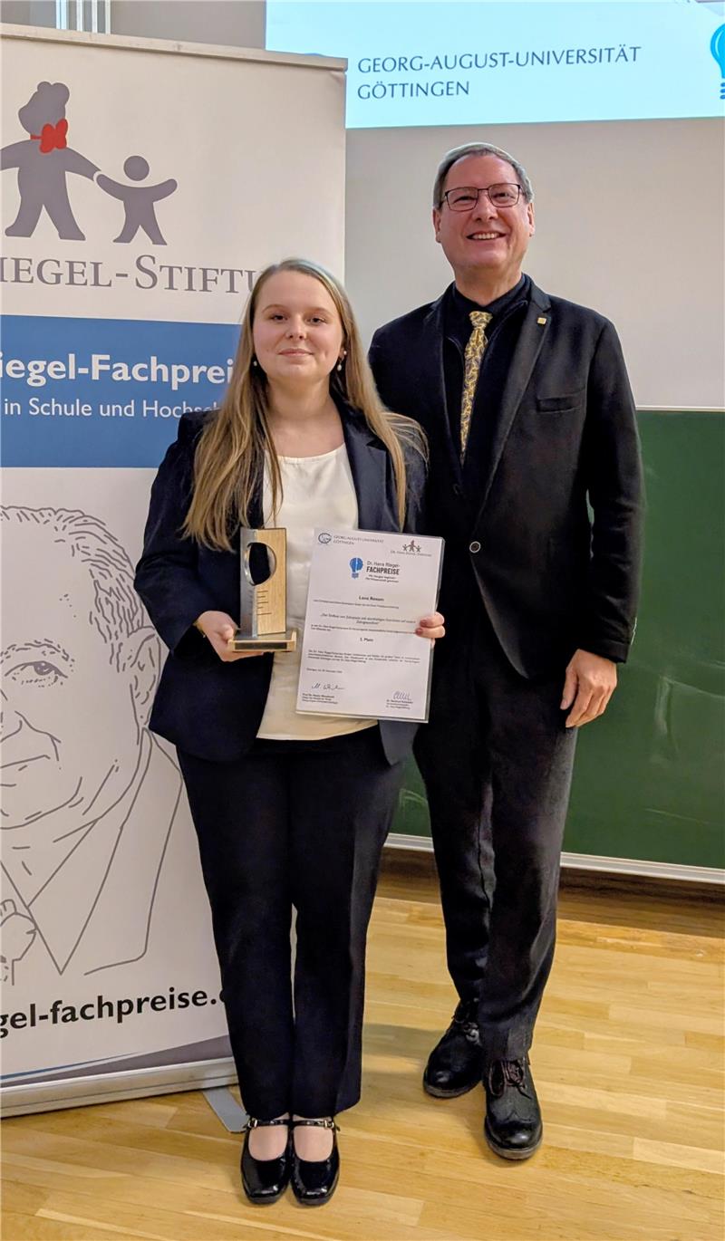 Zwei Personen in formeller Kleidung stehen nebeneinander, eine hält eine Urkunde und eine Auszeichnung, im Hintergrund Banner und Projektionsfläche mit Logos der Georg-August-Universität Göttingen und Dr. Hans Riegel-Fachpreise.