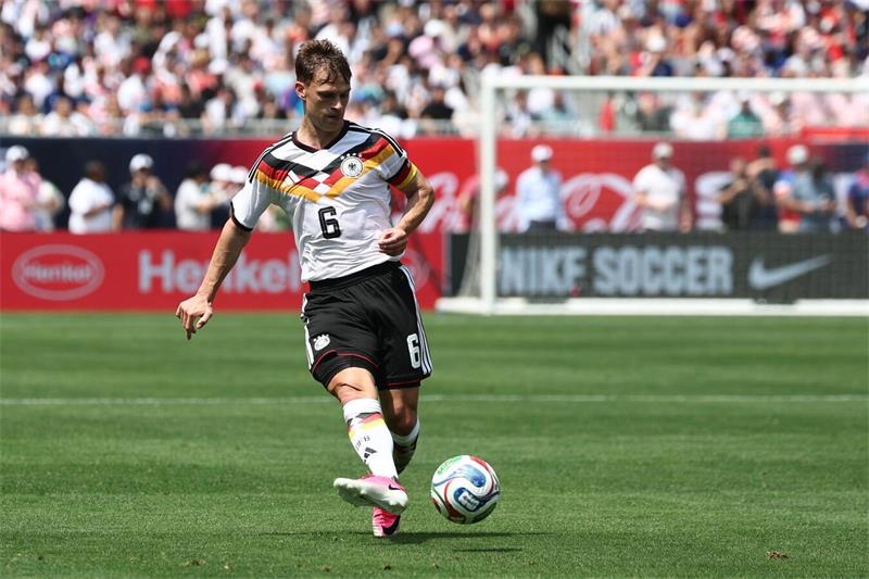 Ein Fußballer trägt ein weißes Trikot auf dem sich ein schwarz-rot-goldener Brustring befindet. Er trägt zudem schwarze Hosen und weiße Stutzen und spielt gerade den Ball.