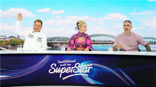 Ein Mann, eine Frau und ein weiterer Mann sitzen hinter einem TV-Pult, im Hintergrund ist die Skyline von Köln zu sehen.