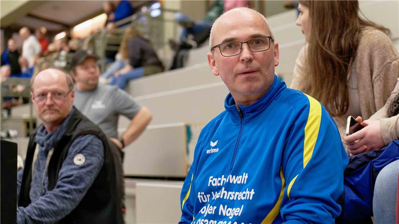 Person in blau-gelbem Trainingsanzug mit Werbeaufdruck sitzt auf Tribüne, weitere Personen im Hintergrund auf Sitzreihen.