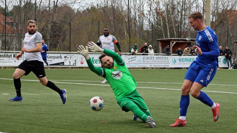 Torwart in grüner Kleidung taucht nach rechts zum Ball auf einem Fußballfeld mit Spielern in blauer und weißer Kleidung