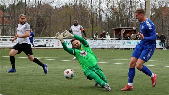 Torwart in grüner Kleidung taucht nach rechts zum Ball auf einem Fußballfeld mit Spielern in blauer und weißer Kleidung