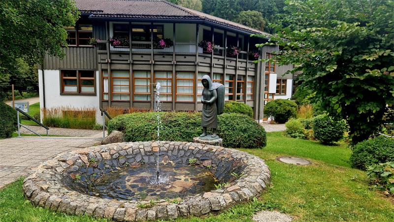 Steinbrunnen mit kleiner Fontäne und Kiepenfrau-Statue vor einem Gebäude mit Blumenkästen.