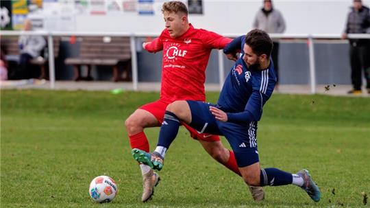 Zwei Fußballspieler kämpfen um den Ball, einer in rotem Trikot, der andere in blauem Trikot, auf einem Rasenplatz.