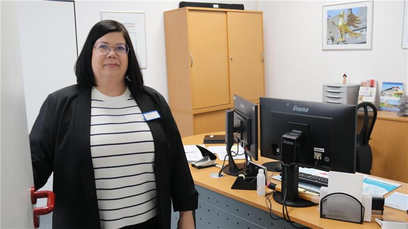 Frau mit schwarzer Jacke und weiß-schwarz gestreiftem Shirt steht in einem Büro mit Schreibtisch, Computer und Aktenschrank im Hintergrund.