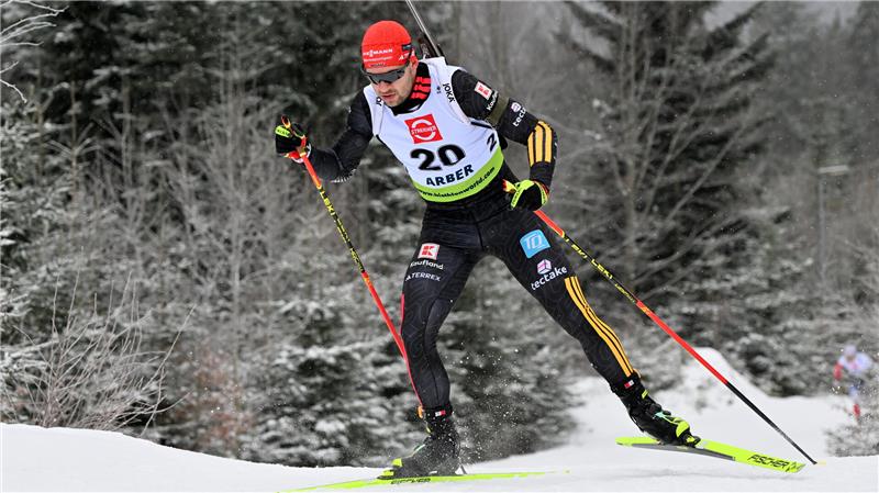 Biathhlet mit Startnummer 20 in schwarzer Sportkleidung und roter Mütze beim Laufen im verschneiten Wald.