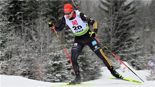 Biathhlet mit Startnummer 20 in schwarzer Sportkleidung und roter Mütze beim Laufen im verschneiten Wald.