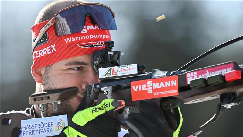 Biathlet mit rotem Stirnband und schwarzer Sportbrille zielt mit einem Gewehr, auf dem mehrere Sponsorenaufkleber sichtbar sind.