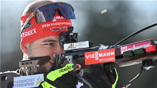 Biathlet mit rotem Stirnband und schwarzer Sportbrille zielt mit einem Gewehr, auf dem mehrere Sponsorenaufkleber sichtbar sind.