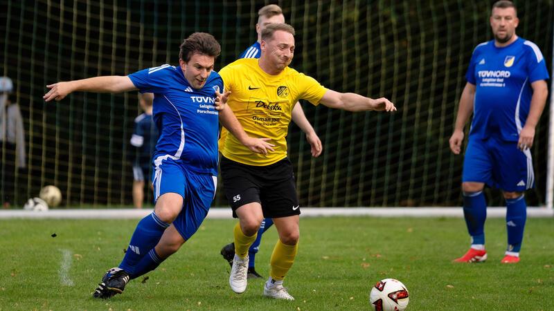 Zwei Fußballspieler in gelbem und blauem Trikot kämpfen um den Ball auf dem Spielfeld, im Hintergrund zwei weitere Spieler und ein Tor