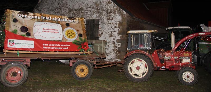 Roter Traktor zieht Anhänger mit beleuchtetem Werbebanner vor altem, weiß gestrichenem Gebäude bei Nacht.