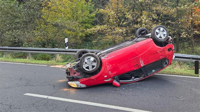 Rotes Auto liegt auf dem Dach auf einer Straße neben einer Leitplanke vor einem Wald.