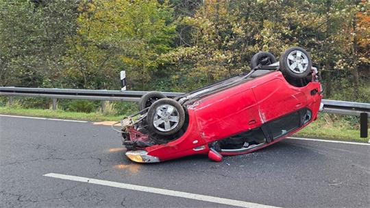 Rotes Auto liegt auf dem Dach auf einer Straße neben einer Leitplanke vor einem Wald.
