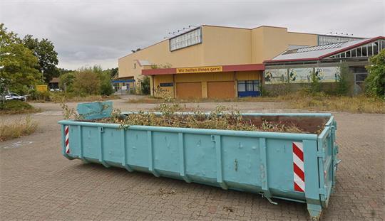 Großer blauer Abfallcontainer auf einem Parkplatz vor einem gelblichen Gebäude mit rotem Dach und der Aufschrift 'Wir helfen Ihnen gern!'