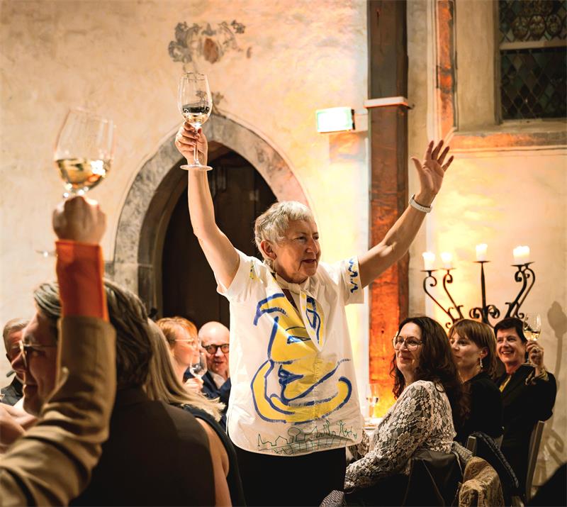 Eine Frau mit einem Glas Wein in der Hand und einer bemalten Bluse streckt inmitten einer Menschenmenge ausgelassen beide Arme in die Höhe.