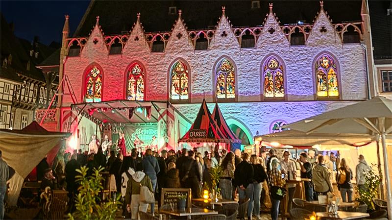 Beleuchtetes historisches Gebäude mit gotischen Fenstern bei Nacht vor einer Menschenmenge auf einem belebten Platz mit Zelten und Tischen