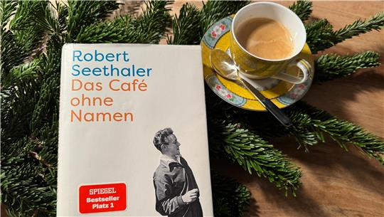 Das Bild zeigt das Buch "Das Café ohne Namen" auf einem Tannenzweig, daneben eine Tasse Kaffee.