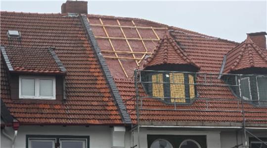 Dach mit roten Ziegeln, teilweise abgedeckt, mit sichtbarem Holzgerüst und Gerüst vor einem Erkerfenster.