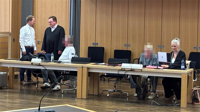 Zwei Männer stehen und sprechen an einem langen Holztisch, zwei Personen sitzen mit verschwommenen Gesichtern, eine Frau arbeitet an einem Laptop