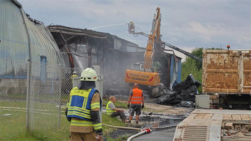 Feuerwehrmann in Schutzkleidung vor einem beschädigten Industriegebäude, in dem ein Bagger mit ausgefahrenem Arm arbeitet und mehrere Personen in Warnwesten am Boden stehen