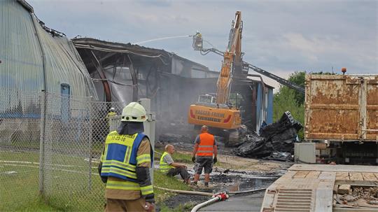 Feuerwehrmann in Schutzkleidung vor einem beschädigten Industriegebäude, in dem ein Bagger mit ausgefahrenem Arm arbeitet und mehrere Personen in Warnwesten am Boden stehen