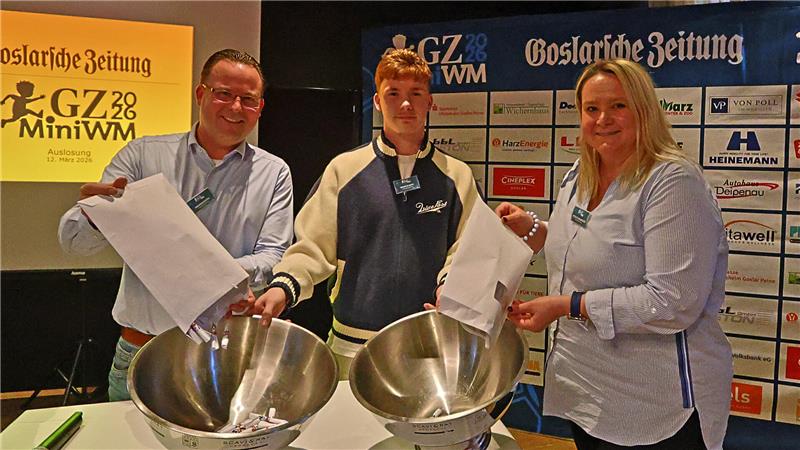Drei Personen stehen an einem Tisch mit zwei großen Metallschüsseln und ziehen Zettel aus weißen Umschlägen vor einem Werbeplakat der Goslarschen Zeitung und Mini-WM-Logos