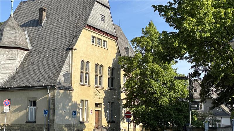 Altbau mit steilem Schieferdach und gelber Fassade neben grünen Bäumen an sonnigem Tag